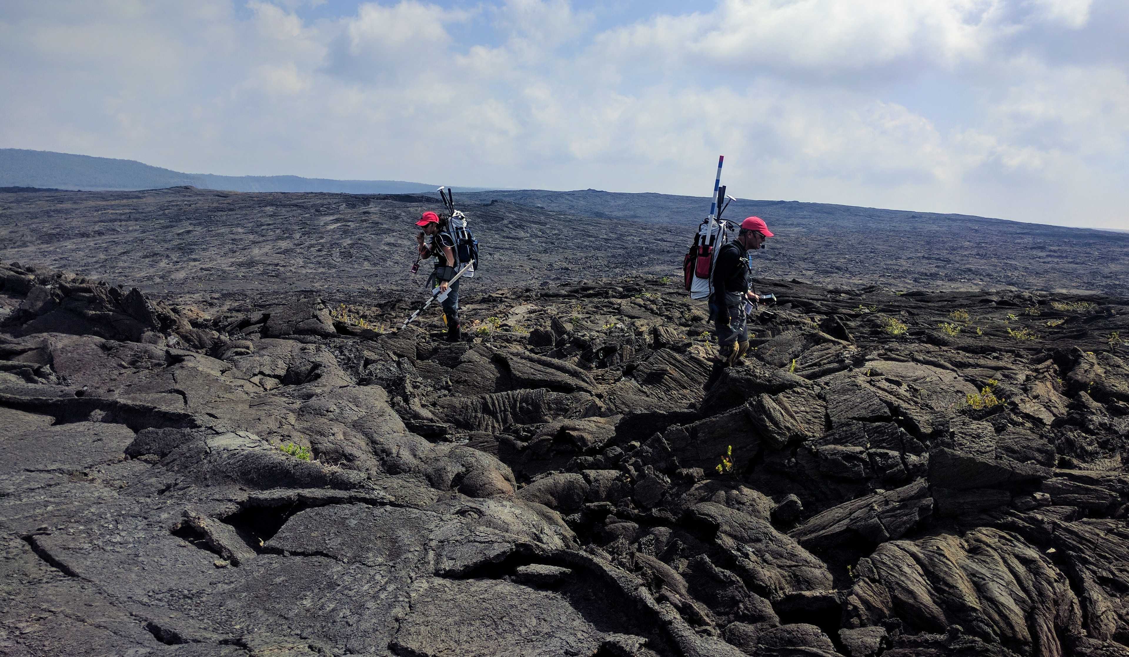 two simulated astronauts walking around on mountainous, basalt-y terrain. they have backpacks with communication equipment and appear to be surveying the surrounding area for potential sample locations